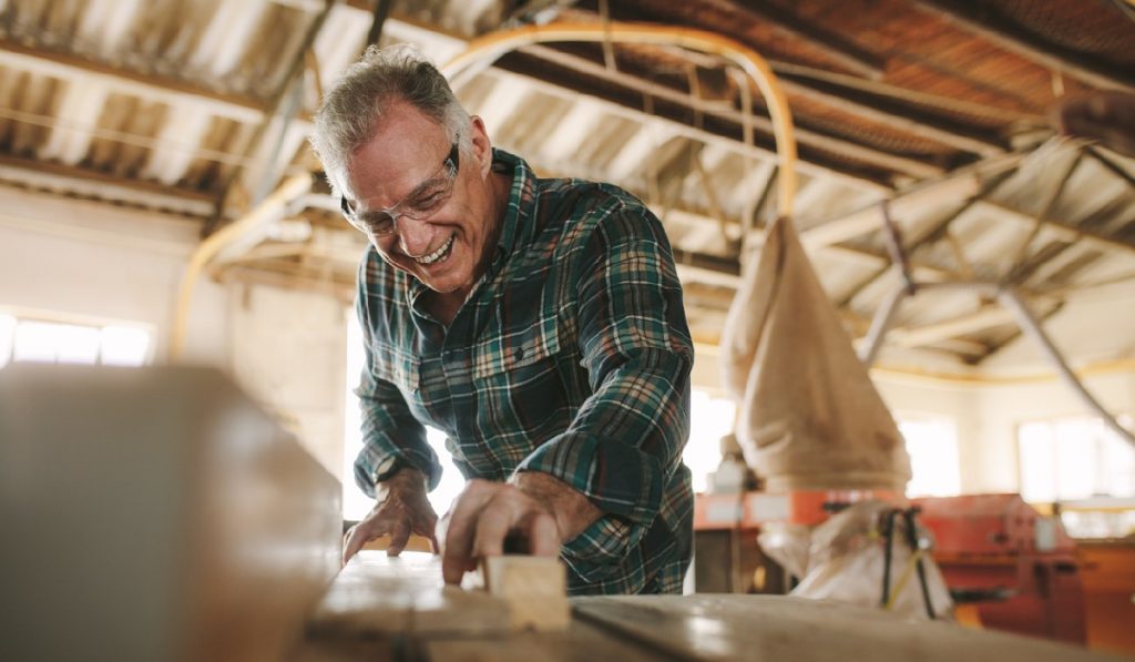 Happy senior male carpenter cutting wood planks on table saw machine. Smiling mature man working in carpentry workshop.