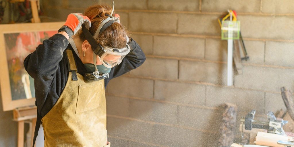 Craftswoman in protective glasses apron and gloves putting on respirator while preparing for work in carpentry workshop