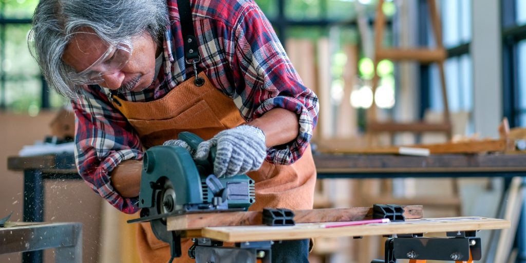 Asian old craftsman use electrical saw to cut wood and generate wood dust in the room with glass windows during day light