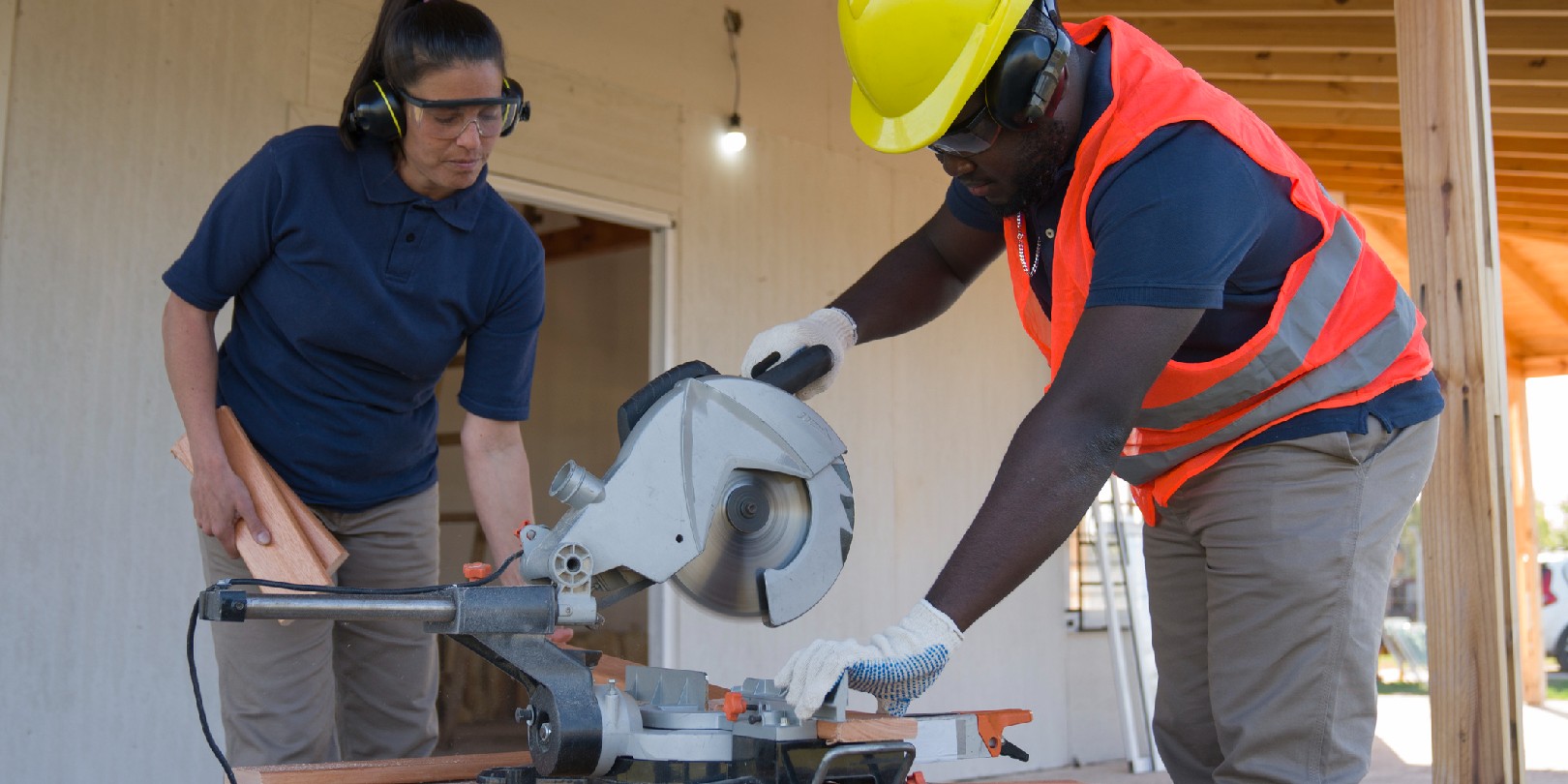 A forewoman is overseeing an afro-american worker sawing a plank during a wood frame house construction