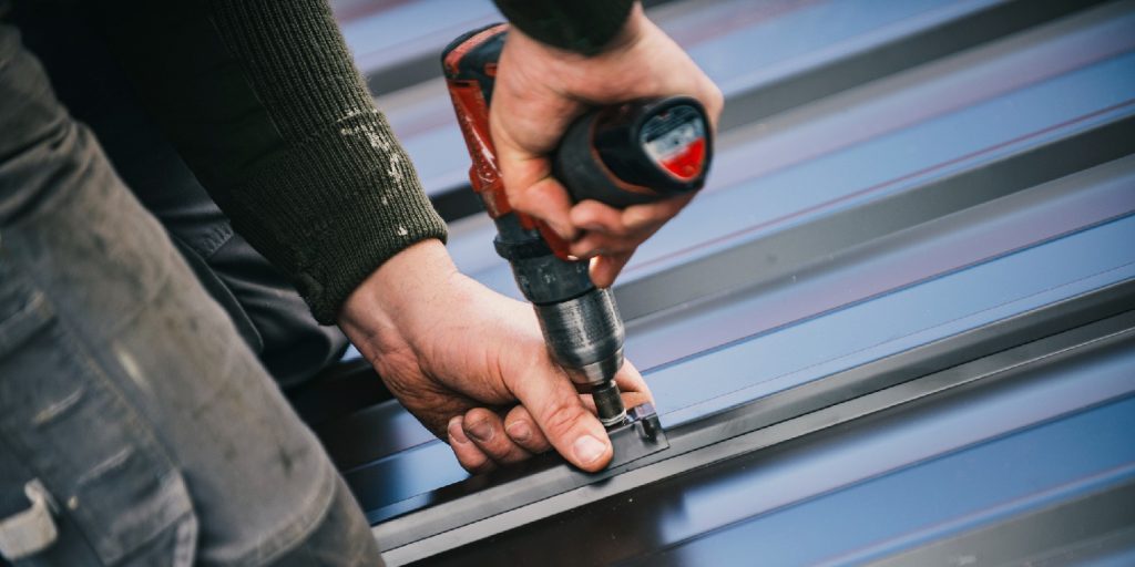 Building contractor is installing metal roofing sheets on the rooftop of the house using electric screwdriver.