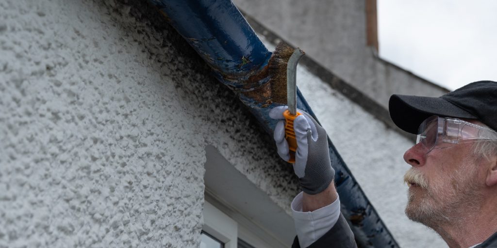 Active retired man using a wire brush to remove old flaking paint from a cast iron roof gutter
