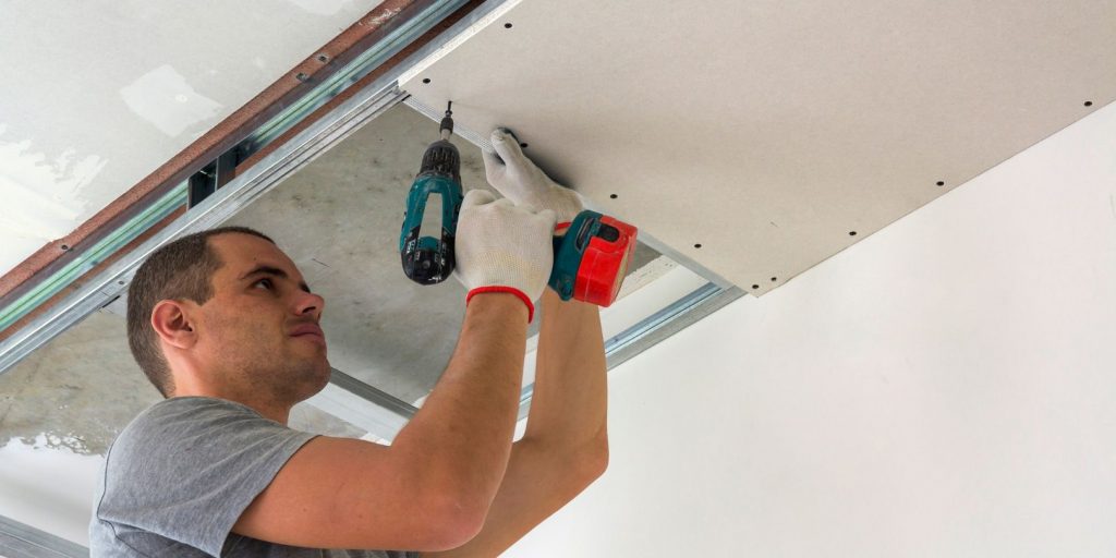 Construction worker assemble a suspended ceiling with drywall and fixing the drywall to the ceiling metal frame with screwdriver.